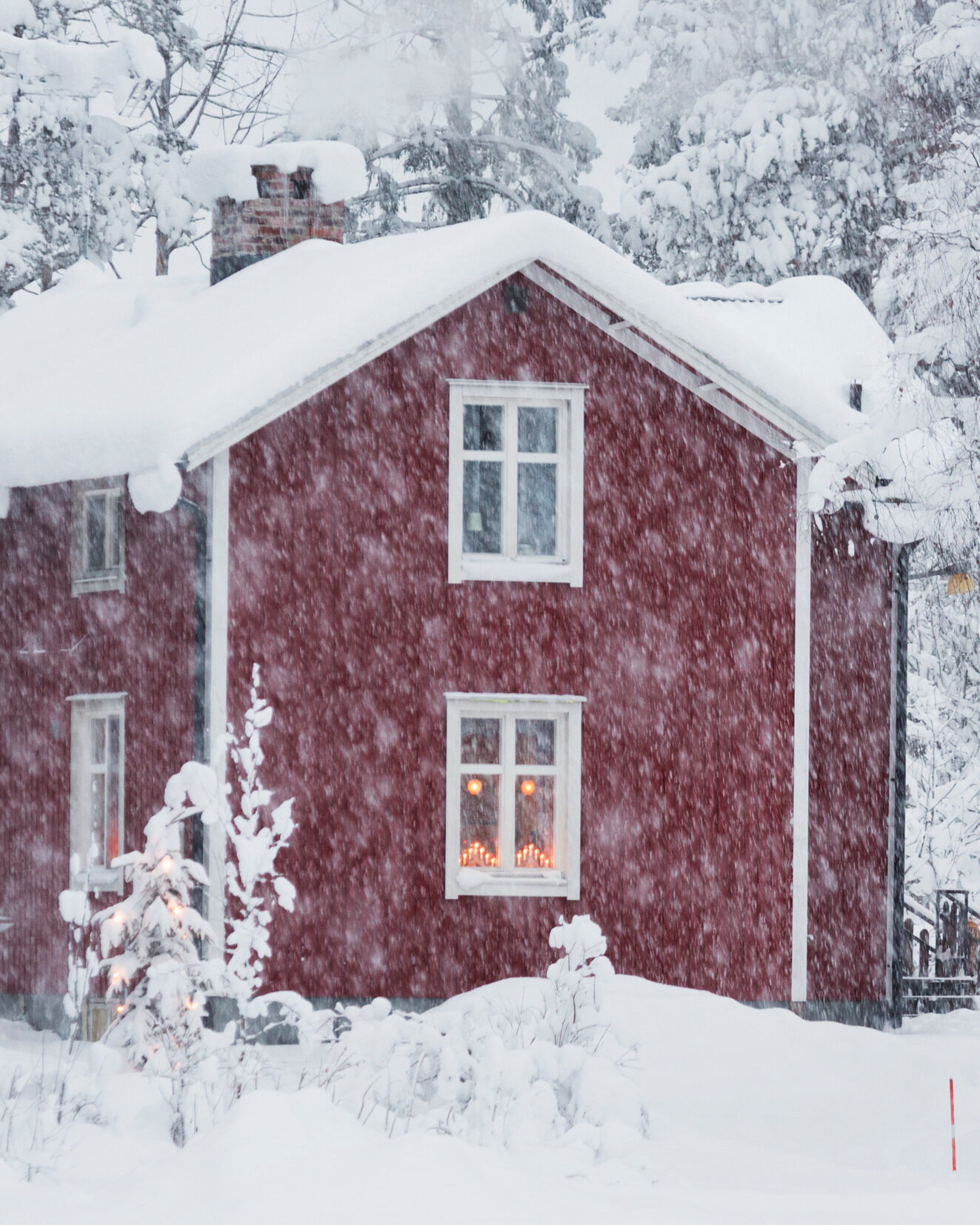 A red house with white trims covered in snow during snowfall.